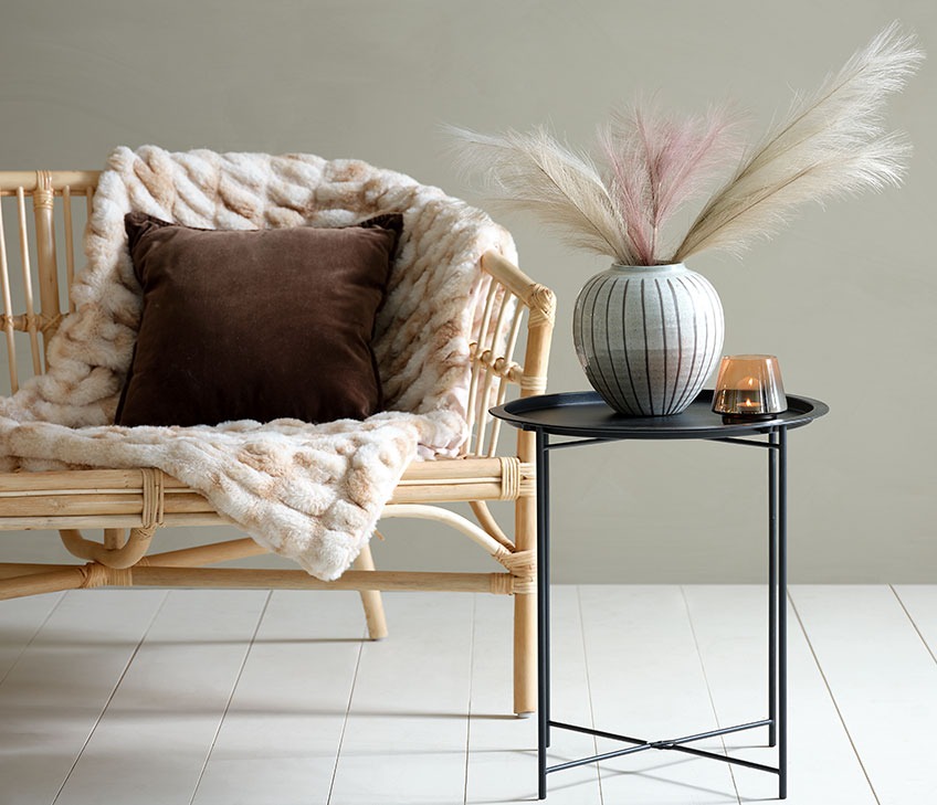 Sofá com manta de pêlo sintético e almofada castanha. Mesa de apoio preta, com vaso e porta-velas